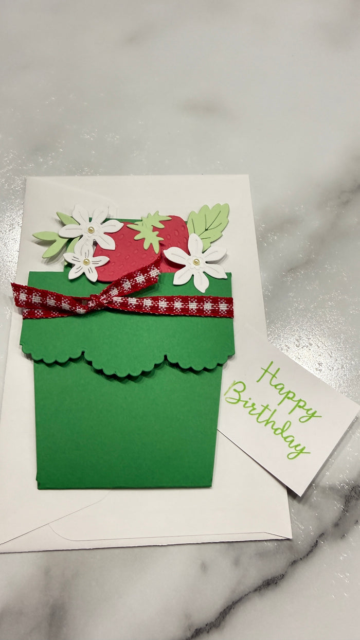 A handmade green flower pot birthday card with a red ribbon, white flowers, and green leaves, shown with a white envelope on a marble surface next to a small 'Happy Birthday' card.