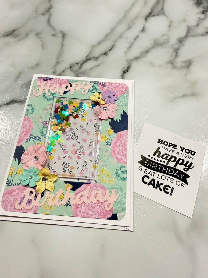 A handmade floral card with a clear shaker window filled with colorful glitter, displayed on a white marble background next to a small white card that reads 'HOPE YOU ARE ALL happy BIRTHS AND EAT LOTS OF CAKE!'.