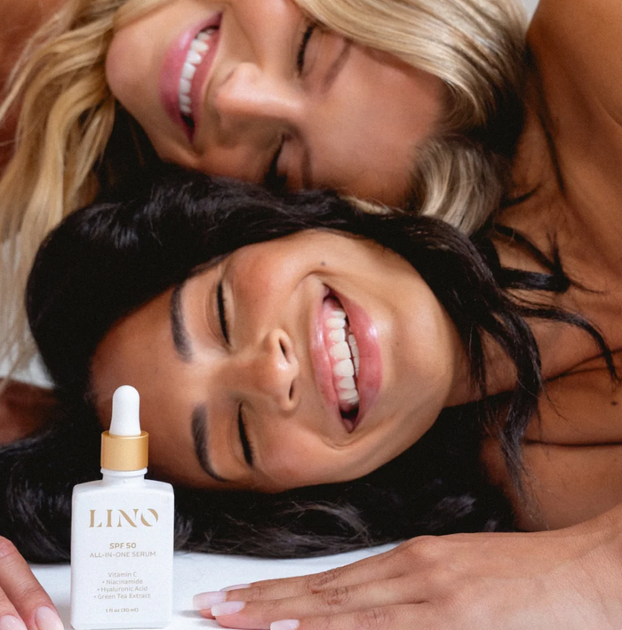 Two smiling women lying close together with a bottle of LINO SPF 50 All-Over Face Serum in the foreground. The product bottle is white with a gold collar and white dropper cap.