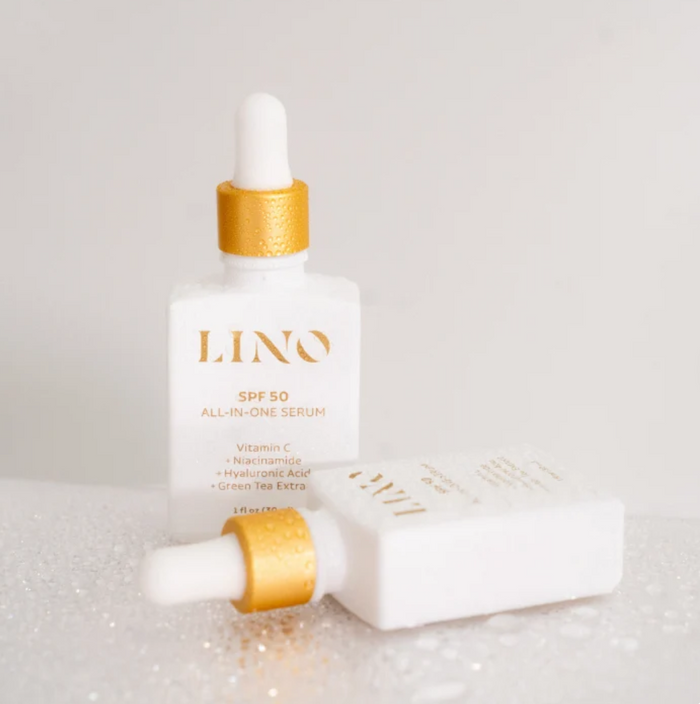 A white skincare bottle with a gold collar and white dropper cap labeled LINO SPF 50, shown with a second bottle lying on its side on a light gray surface with subtle glitter.