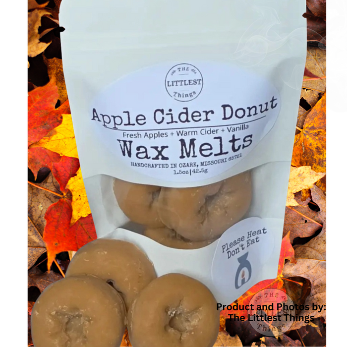 A stand-up pouch labeled 'Apple Cider Donut Wax Melts' by The Littlest Things, shown with several tan donut-shaped wax melts in front, set against a background of colorful autumn leaves.