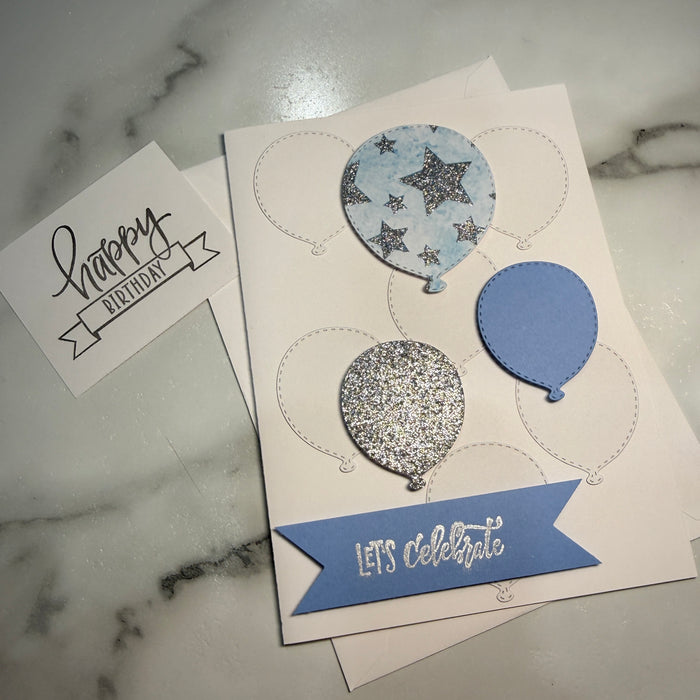 A handmade birthday card with balloon and star cutouts, featuring a blue banner reading 'Let's celebrate' and three balloon shapes in glittery silver and light blue patterns, displayed on a white marble surface with a 'Happy Birthday' card underneath.