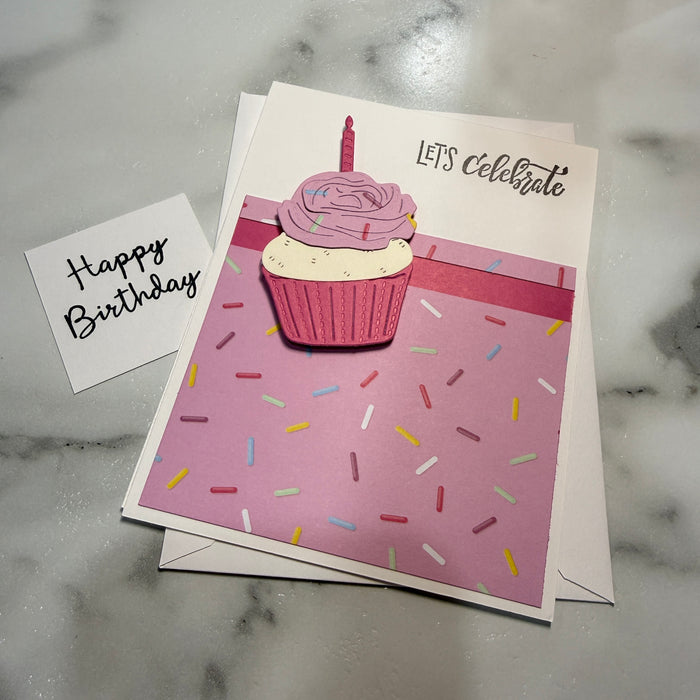 A birthday card set on a light marble surface, featuring a larger square card with a pink cupcake illustration and the words 'LET'S Celebrate' alongside a smaller white card that says 'Happy Birthday' in black script.