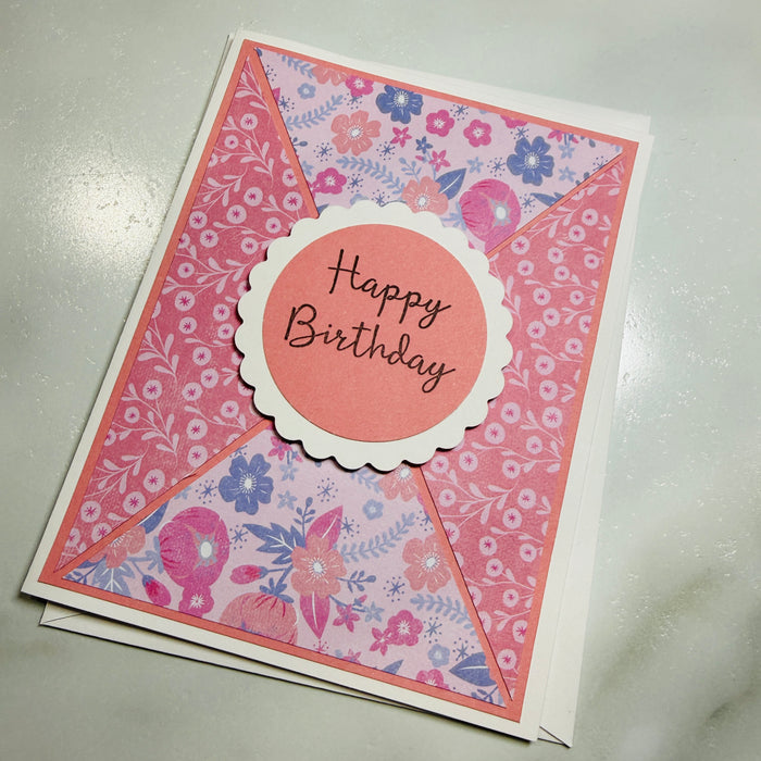 A handmade floral envelope-style birthday card with a scalloped circle reading 'Happy Birthday' in black script, photographed on a light gray surface.