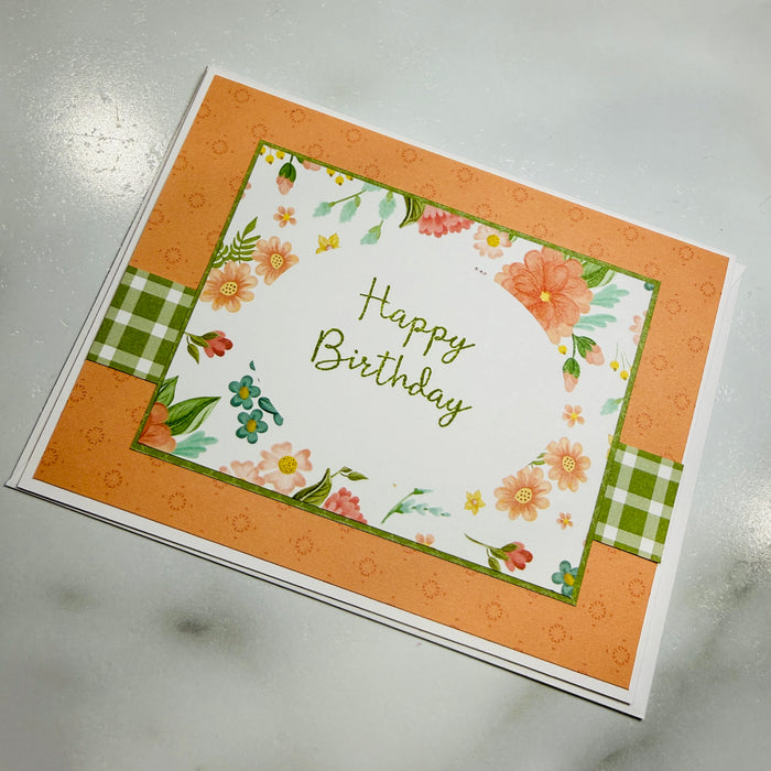 A handmade-style square birthday card with a white center, green and orange floral border, and the words 'Happy Birthday' in green script, photographed on a light marble surface.