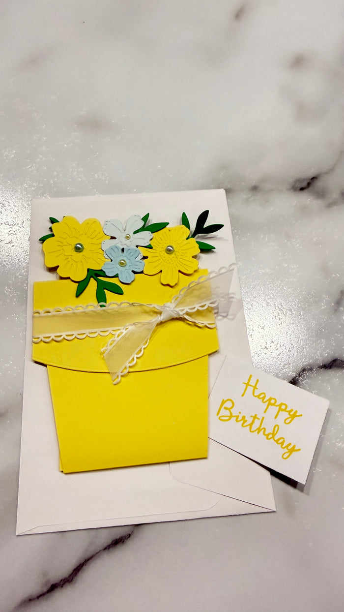 A handmade yellow envelope-style birthday card with layered paper flowers in yellow, light blue, and green, tied with a white lace ribbon, shown on a white marble surface next to a small card reading 'Happy Birthday'.