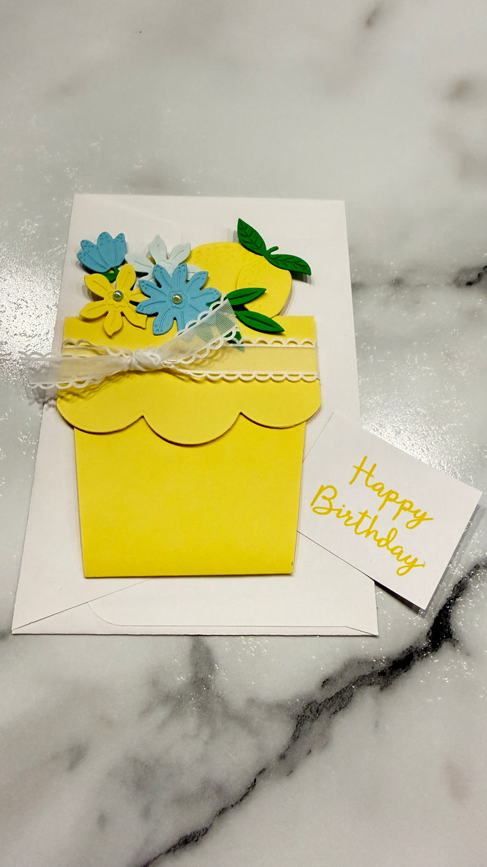A handmade yellow flower pot birthday card with blue and yellow paper flowers, green leaves, and a white lace ribbon, displayed on a white envelope over a marble surface next to a small 'Happy Birthday' card.