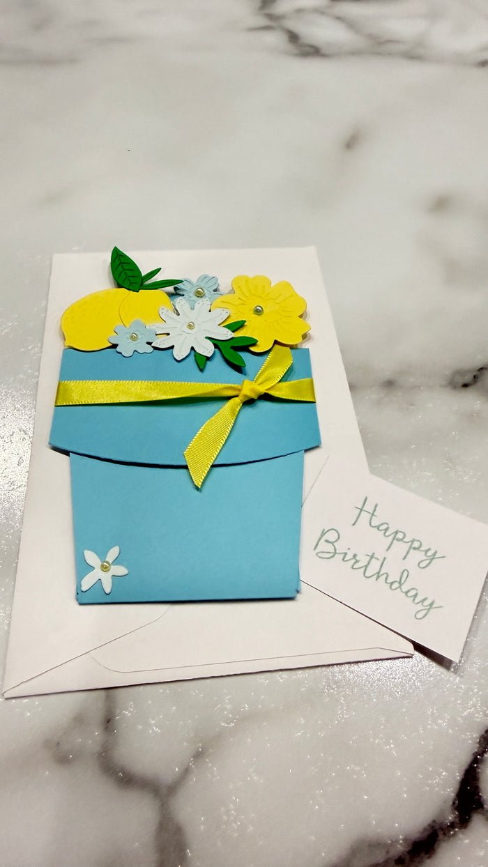 A handmade blue flower pot birthday card with yellow and white paper flowers, a yellow ribbon, and a small white card reading 'Happy Birthday' on a white marble surface.
