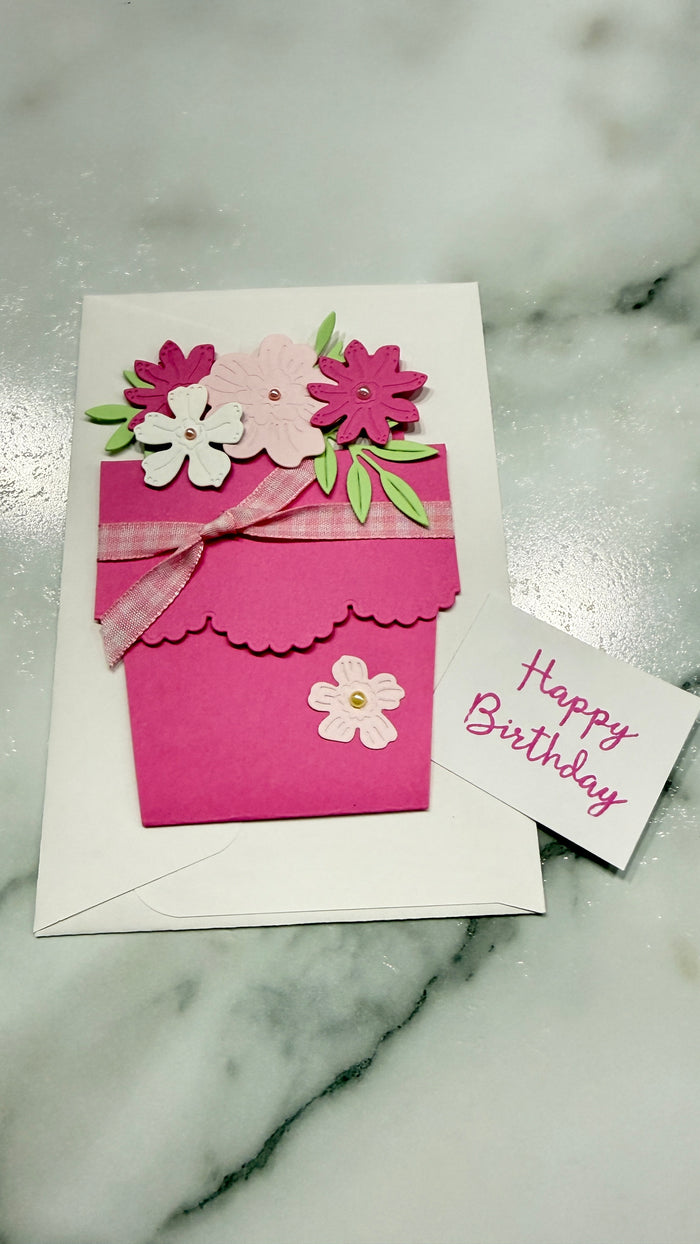 A handmade pink and white birthday card featuring a flower pot design with paper flowers, green leaves, and a pink ribbon, displayed on a white marble surface next to a small white card that reads 'Happy Birthday'.
