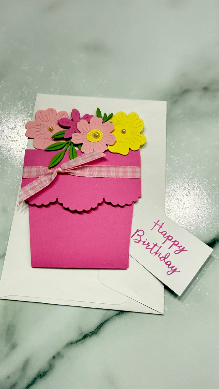 A handmade pink flower pot card with layered paper flowers on top, tied with a pink plaid ribbon, placed on a white envelope over a white marble surface. A small card next to it reads 'Happy Birthday'.