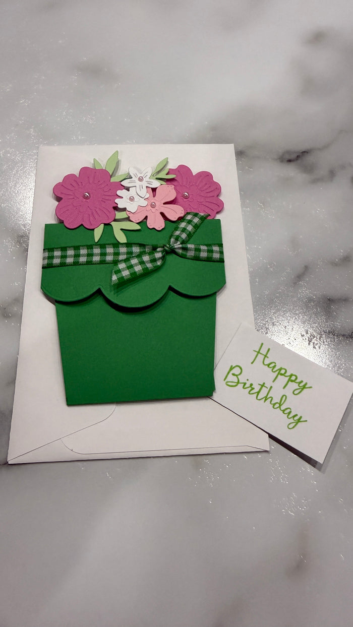 A handmade green pot-shaped birthday card with pink and white paper flowers, a green plaid ribbon, and a small white card reading 'Happy Birthday' on a white envelope, set on a light marble surface.