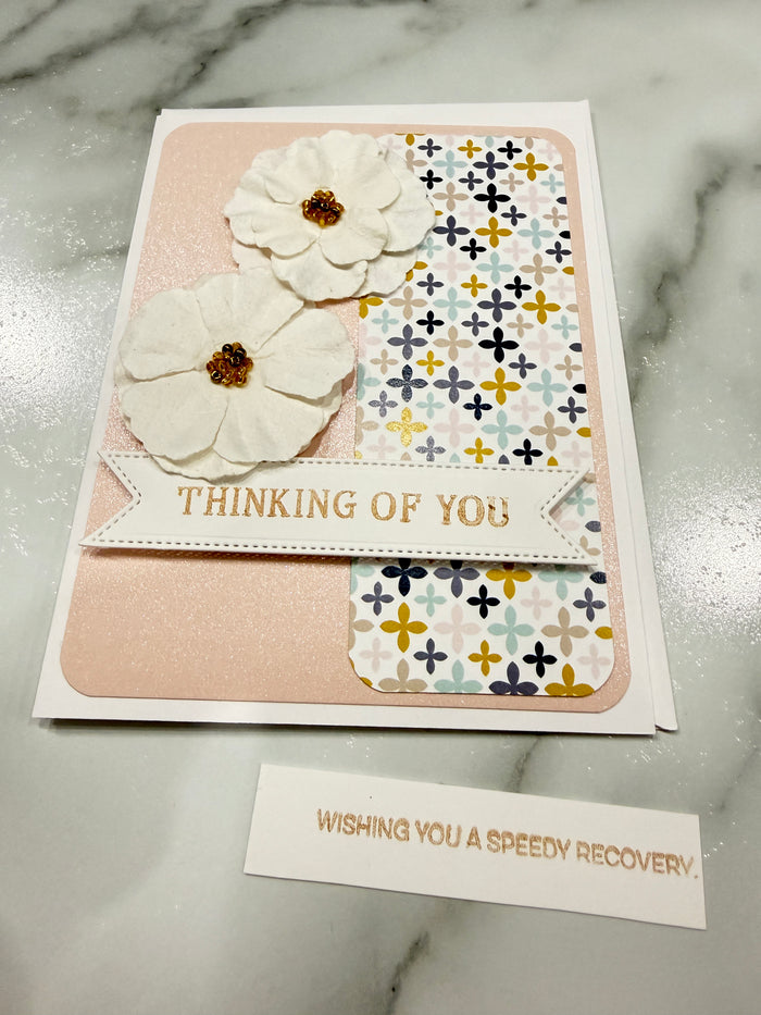 A handmade recovery-themed greeting card with two white paper flowers, a vertical banner reading 'THINKING OF YOU', and patterned paper accents, shown on a white marble surface next to a small card that says 'WISHING YOU A SPEEDY RECOVERY'.