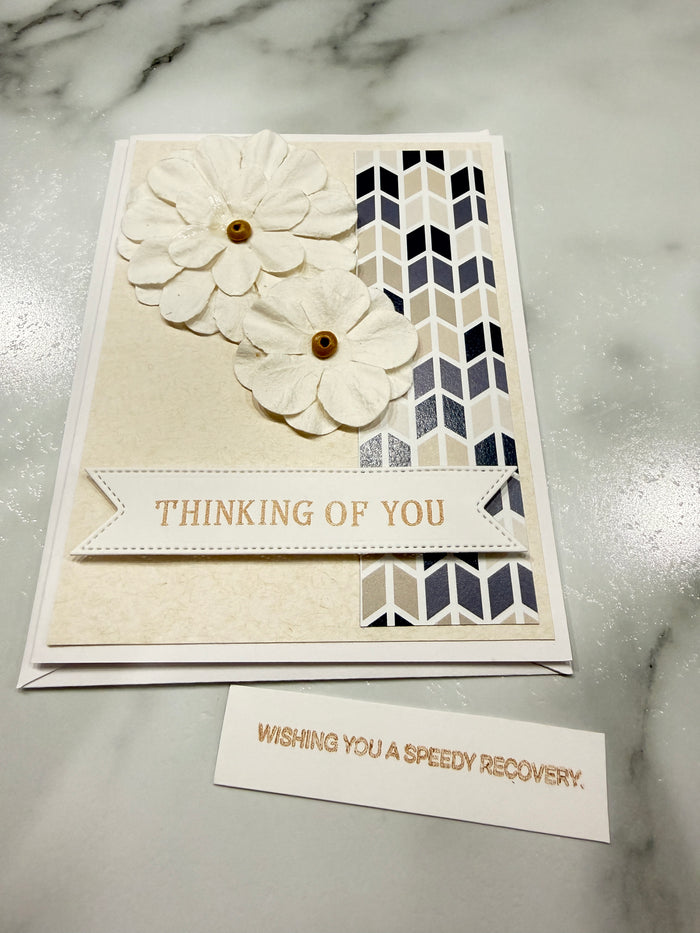 A handmade greeting card with layered white paper flowers, a chevron pattern strip, and a banner reading 'THINKING OF YOU,' shown on a light marble surface with a small card reading 'WISHING YOU A SPEEDY RECOVERY.'.