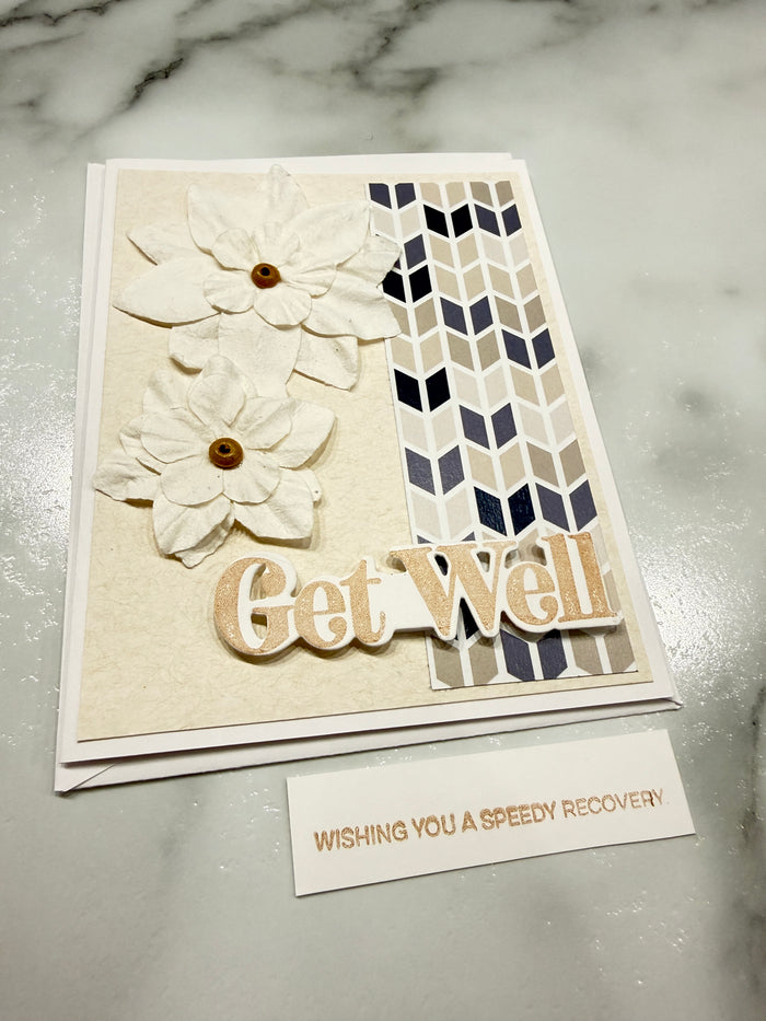 A handmade greeting card with layered cream and beige floral embellishments, a geometric navy, beige, and white chevron pattern, and the words 'Get Well' in raised beige lettering, shown on a light marble surface next to a small card reading 'WISHING YOU A speedy RECOVERY'.