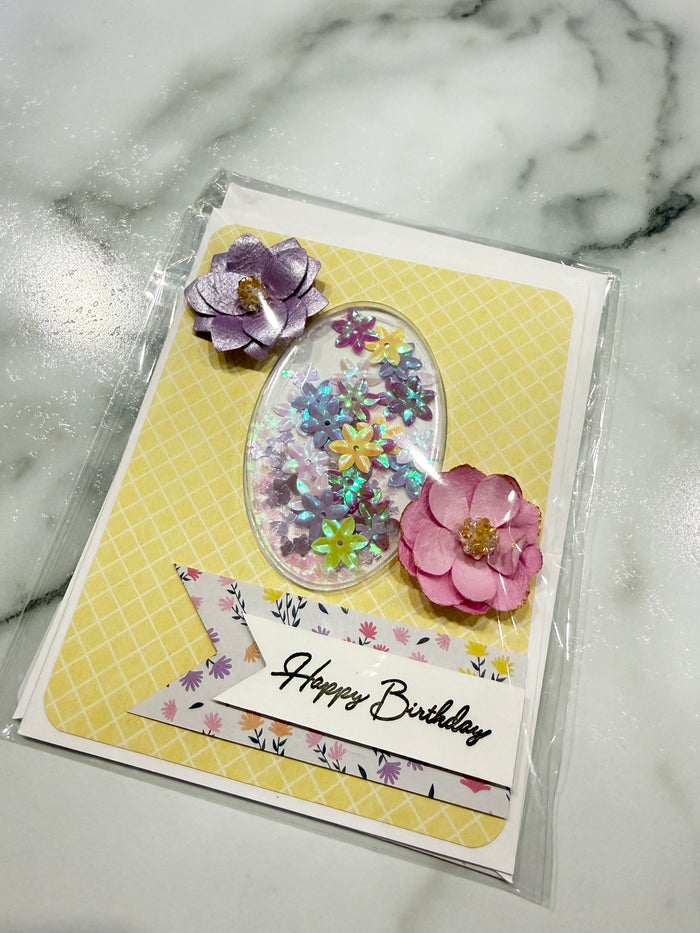 A handmade birthday card in clear plastic packaging, featuring a yellow patterned background, an oval clear resin compartment filled with colorful glitter, pink and purple paper flowers, and a white banner that reads 'Happy Birthday'.