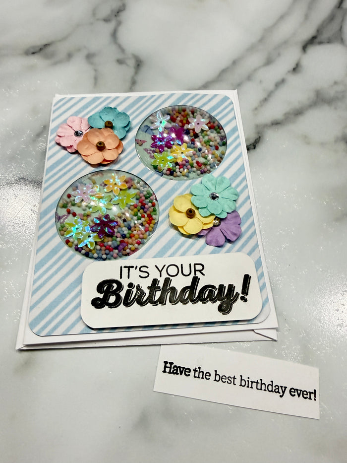 A handmade birthday card with a white and light blue striped background, featuring three circular clear shaker windows filled with multicolored glitter, colorful paper flowers, and a white banner that reads 'IT'S YOUR Birthday!'. A small white tag on the right says 'Have the best birthday ever!'.