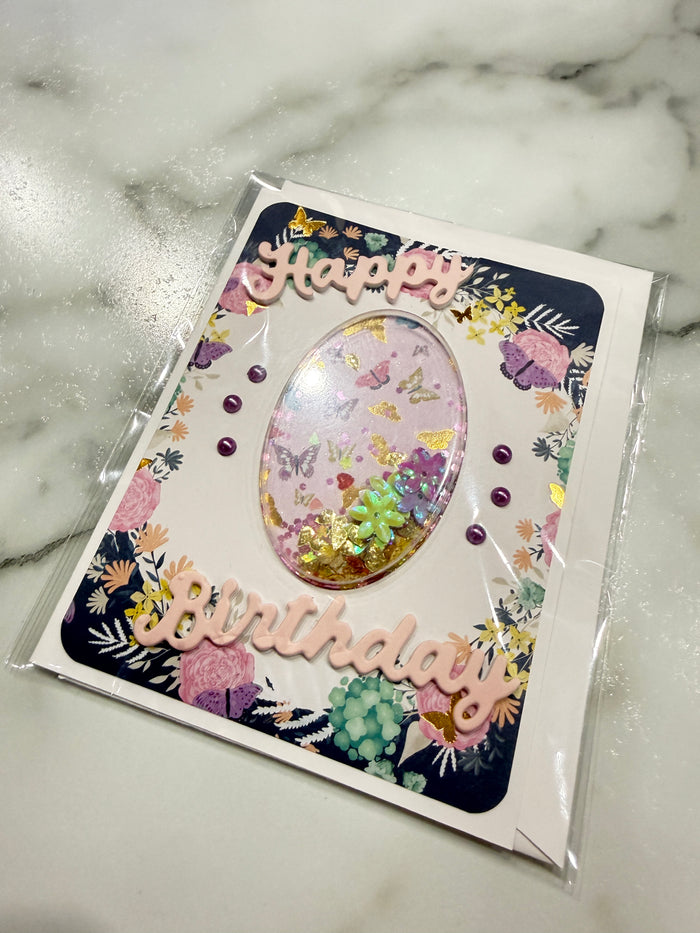 A handmade birthday card inside a clear plastic sleeve, placed on a white marble surface. The card features a white background with a dark navy blue border decorated with colorful floral patterns, 3D pink script text reading 'Happy Birthday', and a central oval window filled with pastel glitter, small flowers, and green leaves.