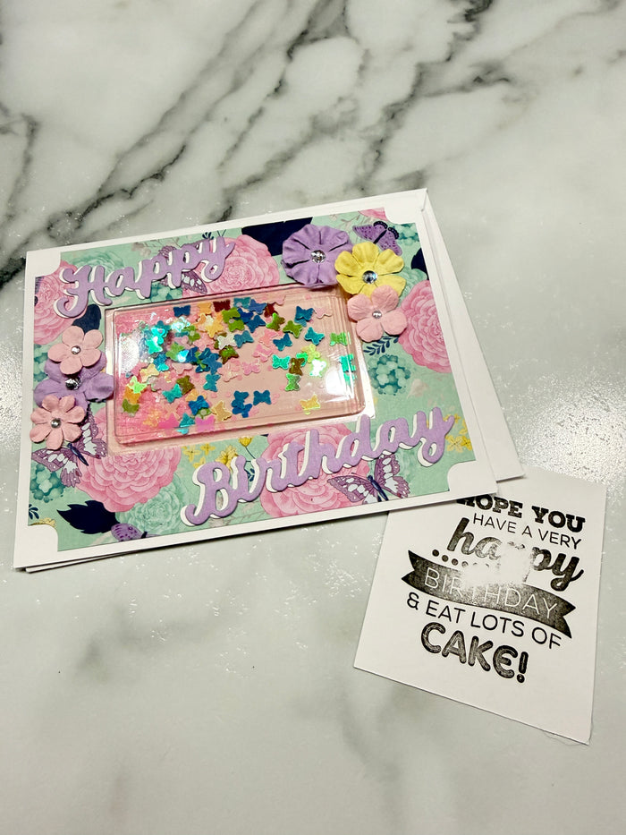 A handmade birthday card with a colorful floral pattern, a clear window filled with glitter, and the word 'BIRTHDAY' in large letters, placed on a white marble surface next to a small printed card.