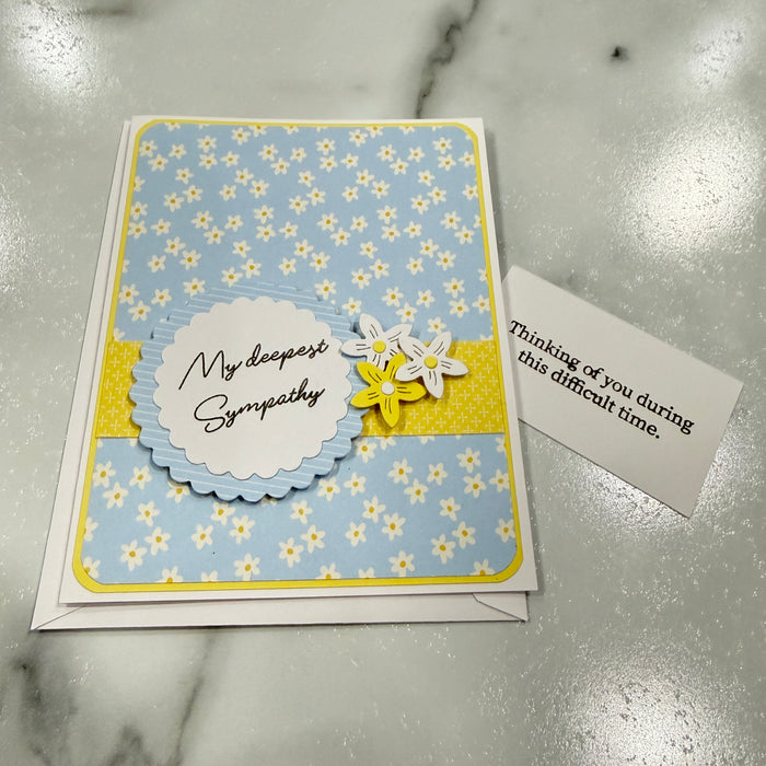 A handmade sympathy card with a light blue and yellow daisy pattern, a scalloped white label reading 'My deepest Sympathy,' and two small layered flowers, shown next to a small note on a marble surface.