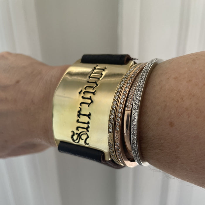 A gold-plated bracelet with the word 'Survivor' engraved on it, worn on a leather strap, displayed on a person's wrist.